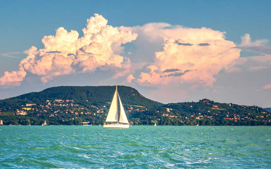 Sehenswuerdigkeiten am Balaton in Ungarn weisses Sightseeing Bootsausflug, Segelboot auf tuerkisblauem See vor gruenen Huegeln und Sommerwolken